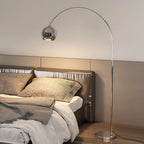 Chrome arc floor lamp from Clérachic arching above a bed with a leather headboard and grey linen, its warm light on the pillows.