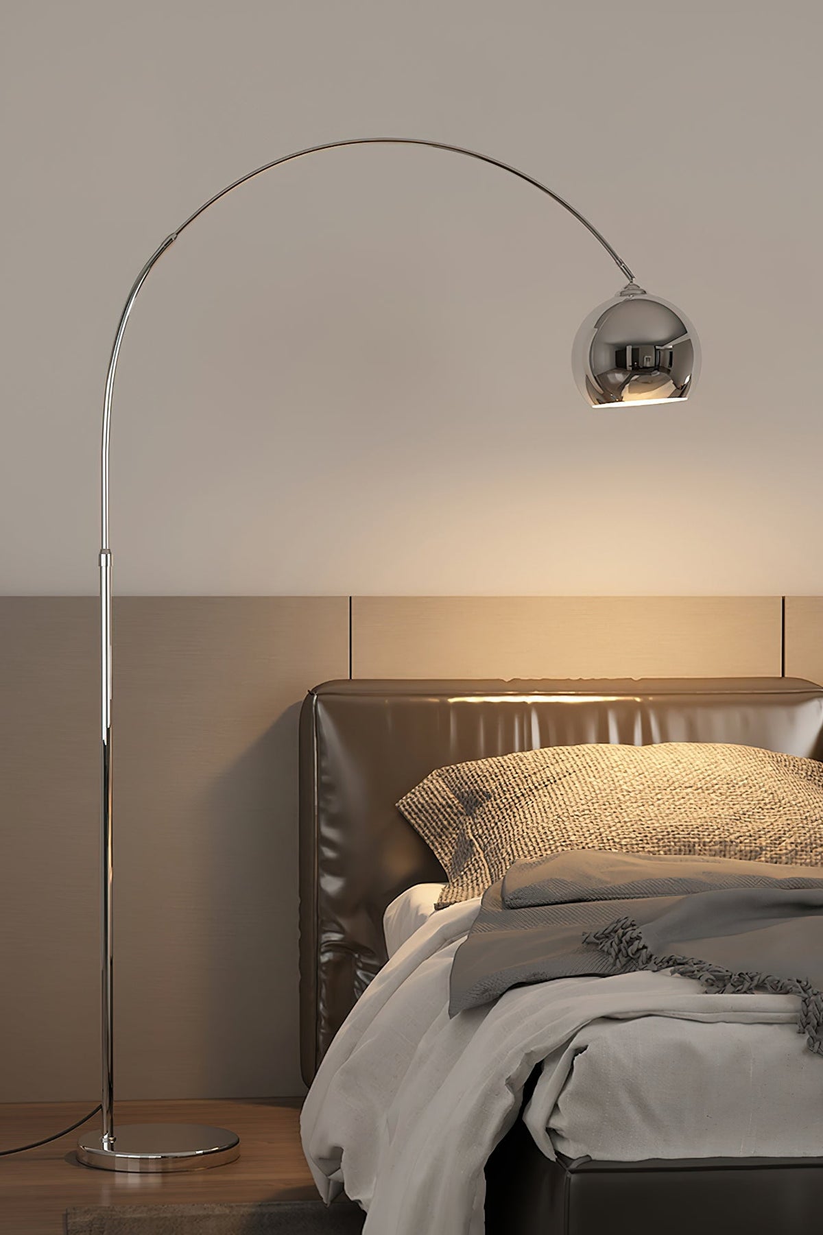 Chrome arc floor lamp from Clérachic standing beside a leather-headed bed, its round head glowing above grey bedding.