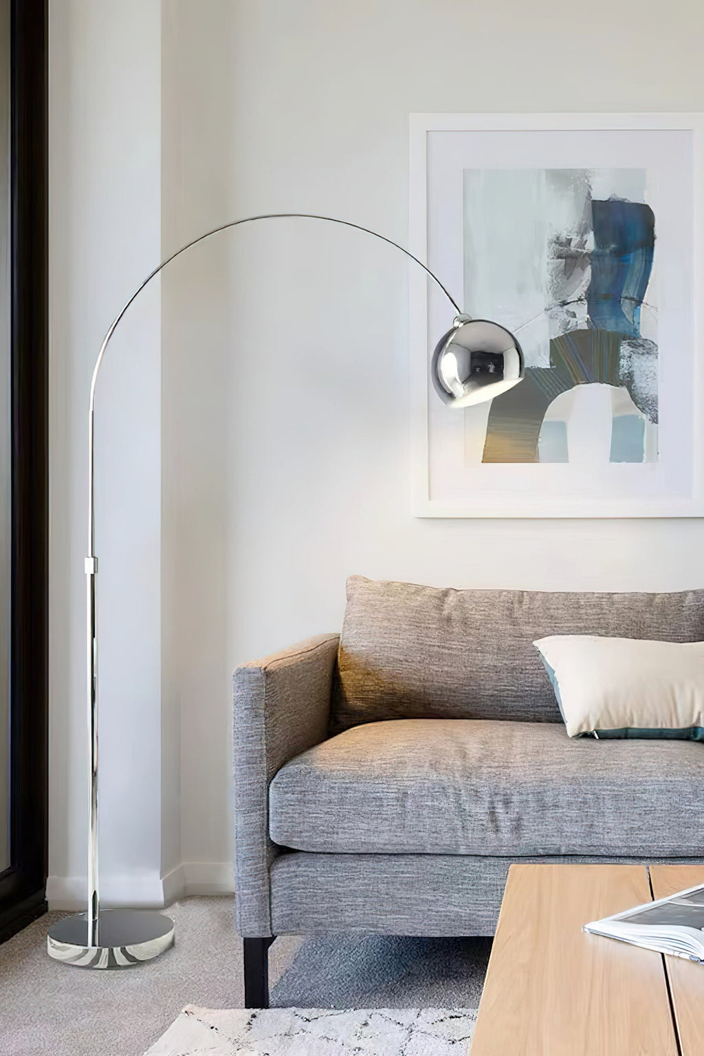 Tall chrome arc lamp from Clérachic over a grey sofa, with an abstract blue and cream painting on the wall behind.