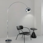 Chrome arc floor lamp from Clérachic reaching over a black chair and small side table in a bare, pale grey room.