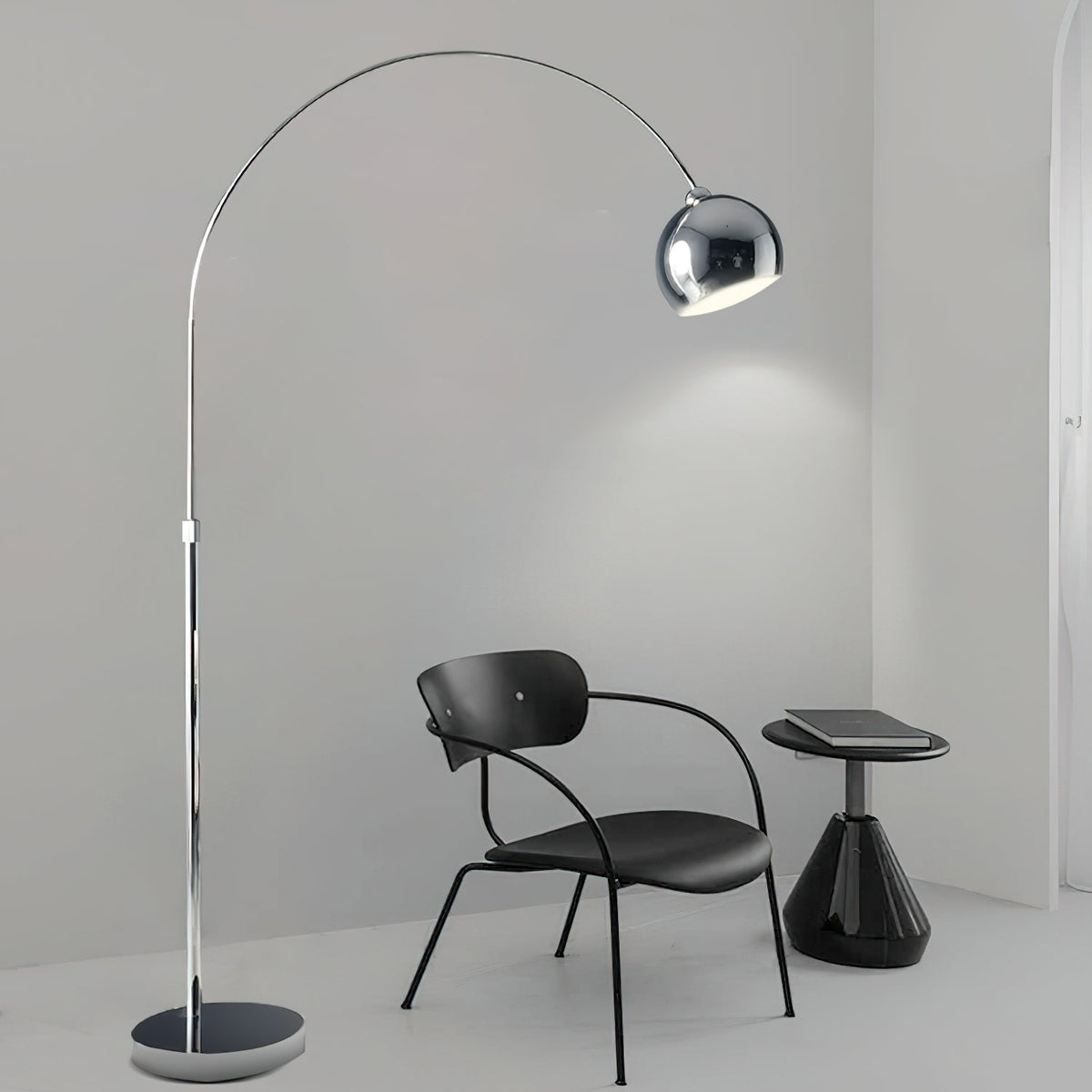 Chrome arc floor lamp from Clérachic reaching over a black chair and small side table in a bare, pale grey room.
