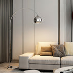 This Clérachic arc lamp's chrome ball head hangs above a cream sofa with taupe cushions, in front of grey curtains.