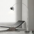 A Clérachic arc lamp curves over a dark lounger with a grey throw, in front of white vertical blinds.