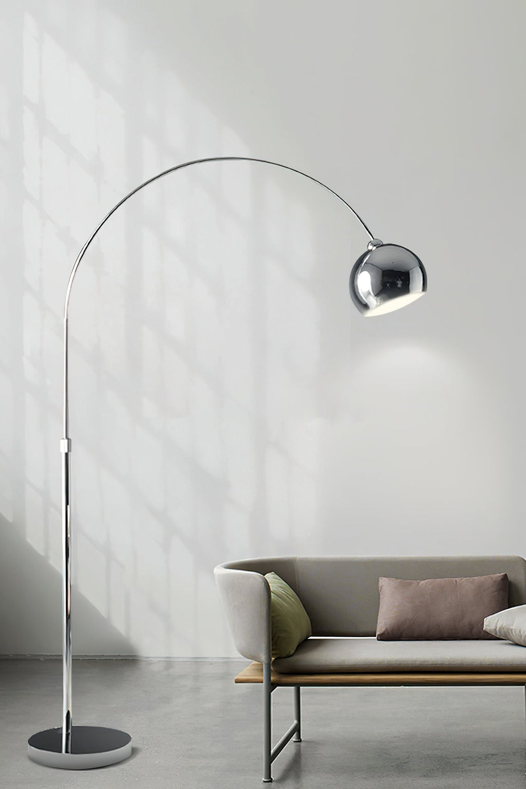 Chrome arc lamp from Clérachic standing in front of a pale wall, with a low sofa and tan cushions in the lower corner.