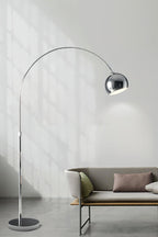 Chrome arc lamp from Clérachic standing in front of a pale wall, with a low sofa and tan cushions in the lower corner.