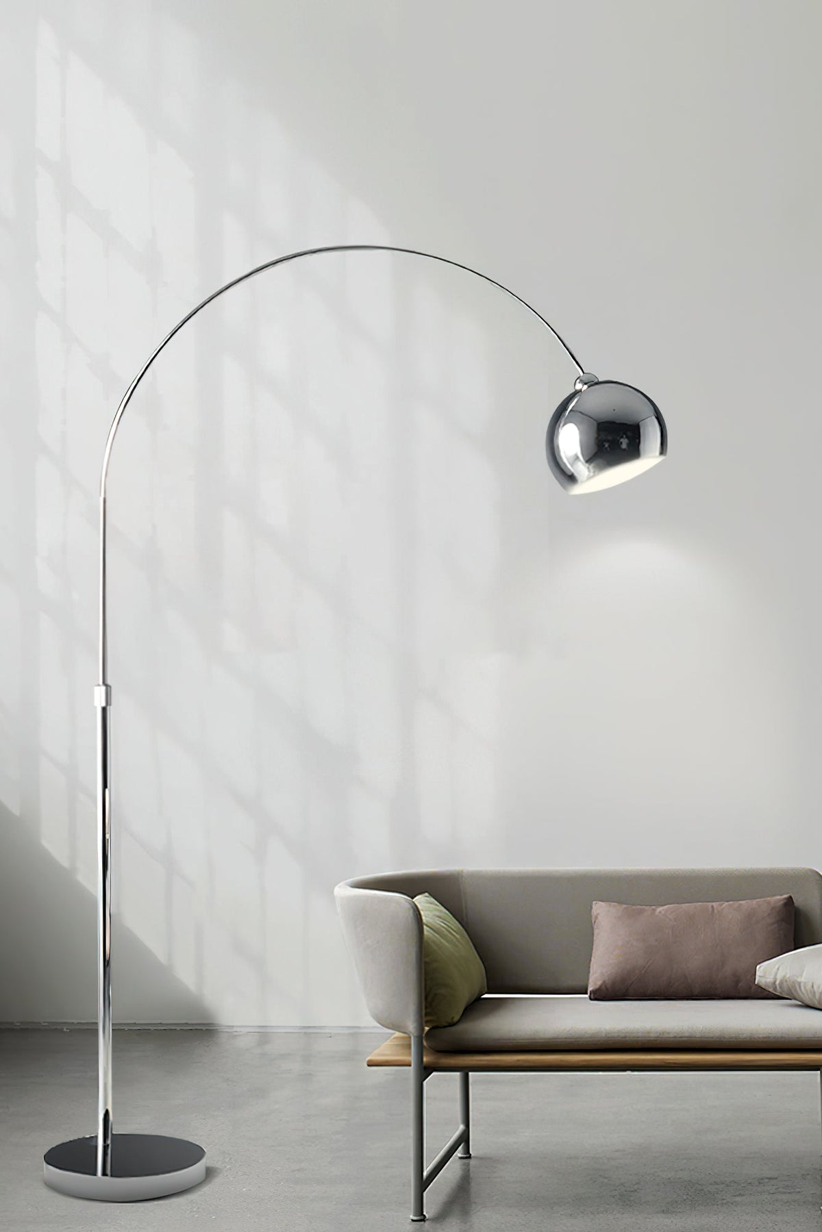 Chrome arc lamp from Clérachic standing in front of a pale wall, with a low sofa and tan cushions in the lower corner.