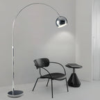 A Clérachic chrome arc lamp arches over a black chair and round side table in a plain grey room.