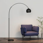 Black arc floor lamp from Clérachic with a matte round head, over a blue armchair and a leafy plant on a wooden floor.