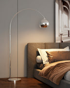 A Clérachic chrome arc lamp leans over a bed with brown bedding and an upholstered headboard, beside an arched wall print.
