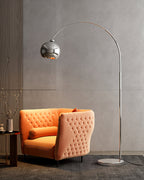 A Clérachic chrome arc lamp curves over an orange tufted armchair, in a corner with a grey wall and a small dark side table.