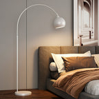 White arc floor lamp from Clérachic with a matte round head and a white base, beside a bed with brown bedding.