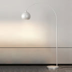 A white Clérachic arc floor lamp with a matte round head and a white base casts light on a pale wall in a minimal room.