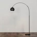 Black arc floor lamp from Clérachic with a matte round head and a black base, on a wooden floor in front of a pale wall.