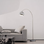 Chrome arc lamp from Clérachic with its head over a grey sofa and a marble table, in a bright room with a curved white wall.