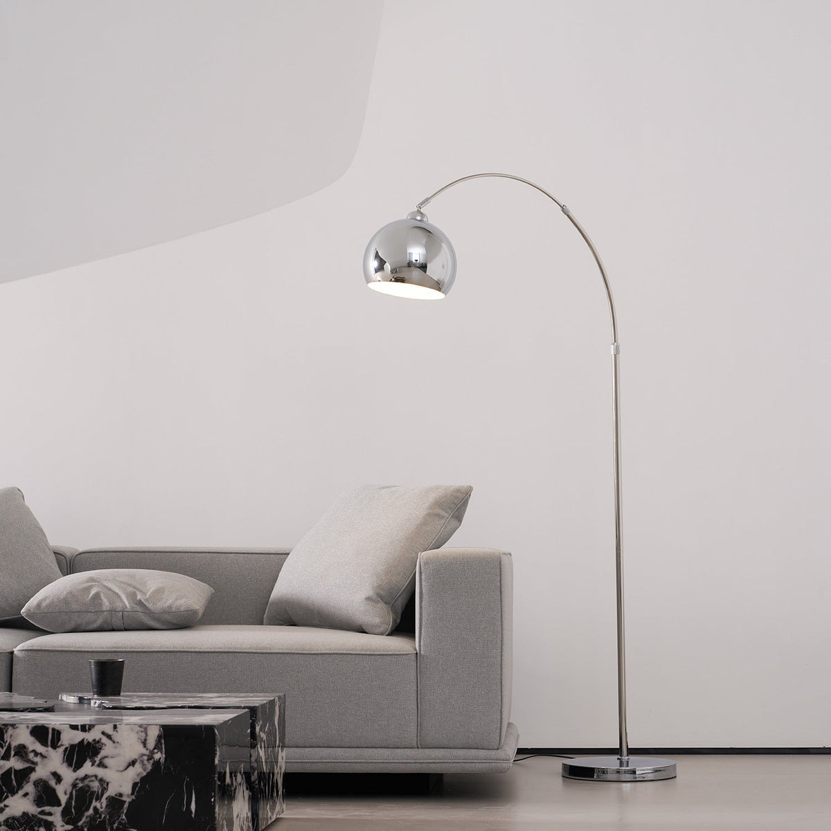 Chrome arc lamp from Clérachic with its head over a grey sofa and a marble table, in a bright room with a curved white wall.