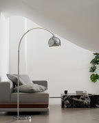 A Clérachic chrome arc lamp rises behind a grey sofa with a wooden plinth, beside a marble table and a monstera plant.