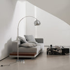 Chrome arc floor lamp from Clérachic with its black cord and in-line foot switch on the floor, beside a grey sofa.