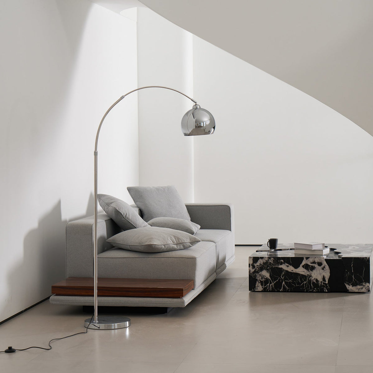 Chrome arc floor lamp from Clérachic with its black cord and in-line foot switch on the floor, beside a grey sofa.