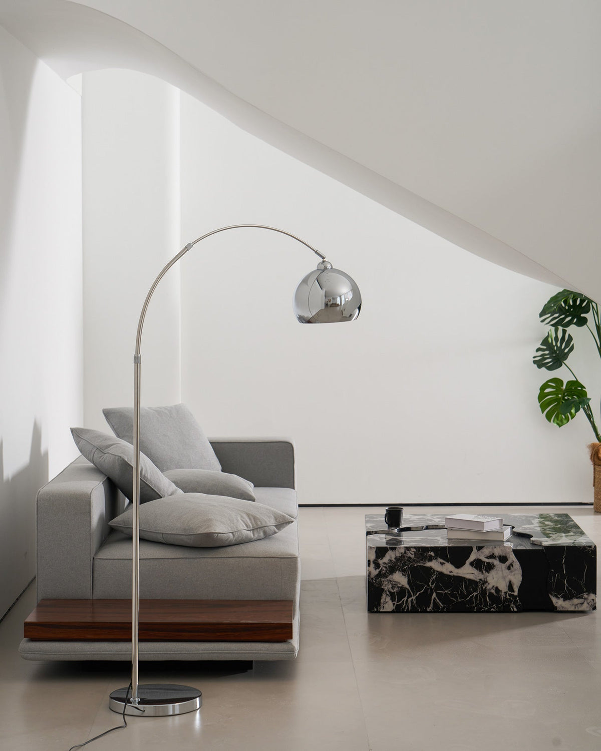 A Clérachic chrome arc lamp leans over a grey sofa with soft cushions, a marble table and a green plant beside it.