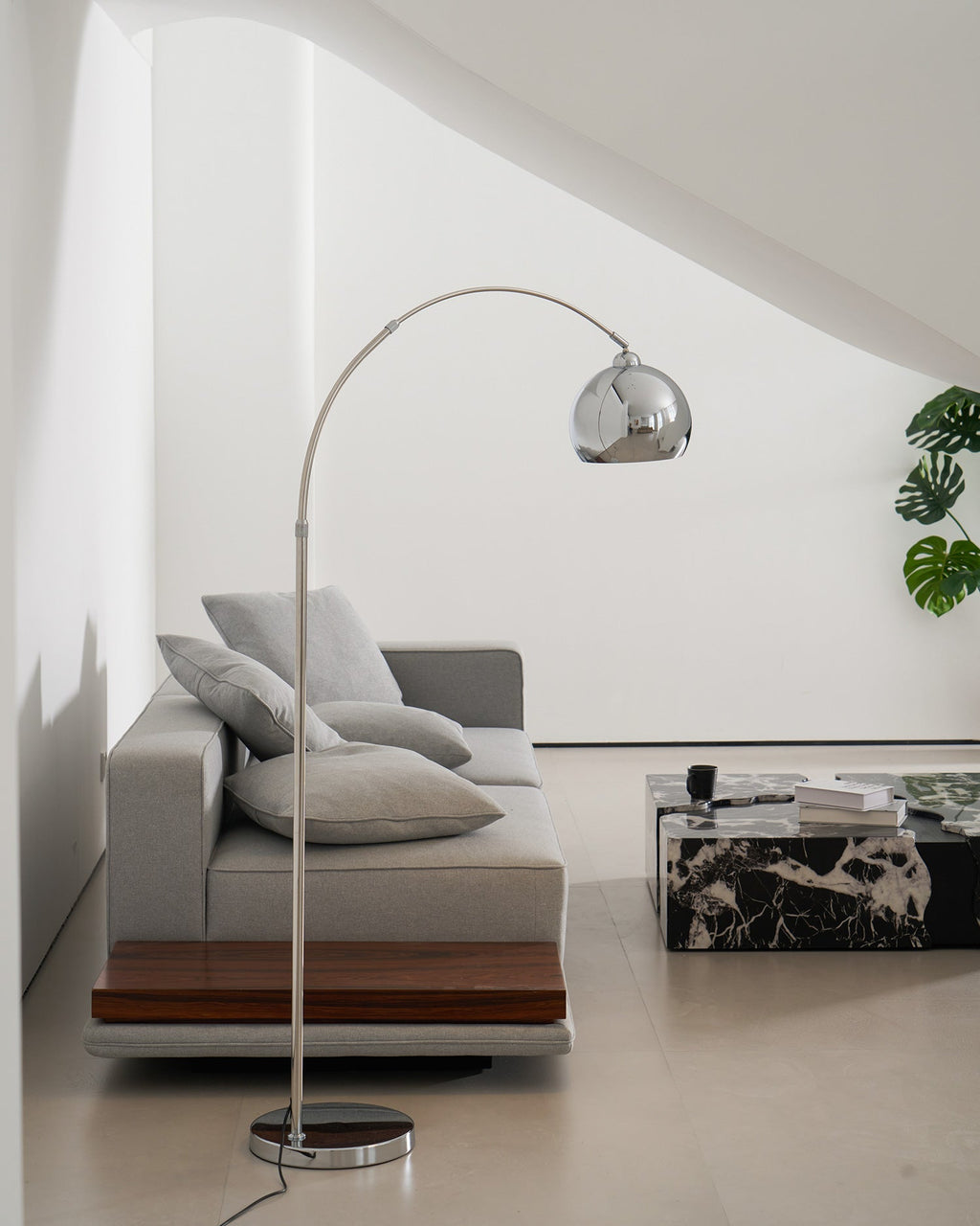 Chrome arc floor lamp from Clérachic standing by a grey sofa with a walnut plinth, close to a dark marble table.