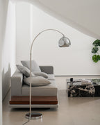 Chrome arc floor lamp from Clérachic standing by a grey sofa with a walnut plinth, close to a dark marble table.