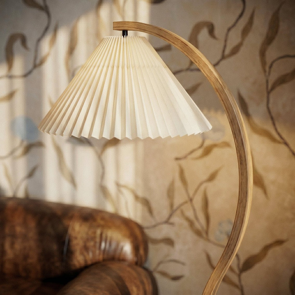 Clérachic floor lamp's cream pleated shade and curved wooden stem, lit in warm light beside a leather sofa arm.