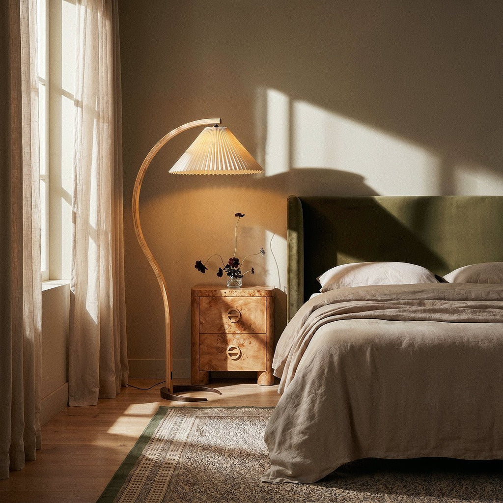 Clérachic arched floor lamp lit beside a burr-wood bedside chest and a linen-dressed bed, with sunlight through sheer curtains.