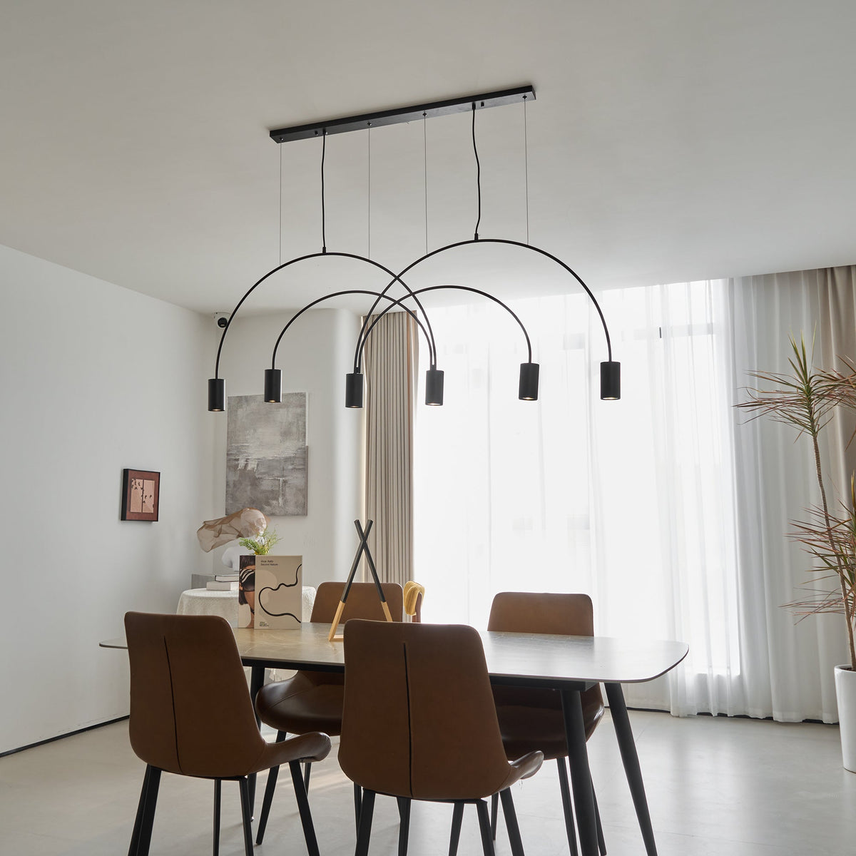 Seen from below, this black Clérachic arch pendant hangs over a dining table with brown chairs and a tall plant by the window.
