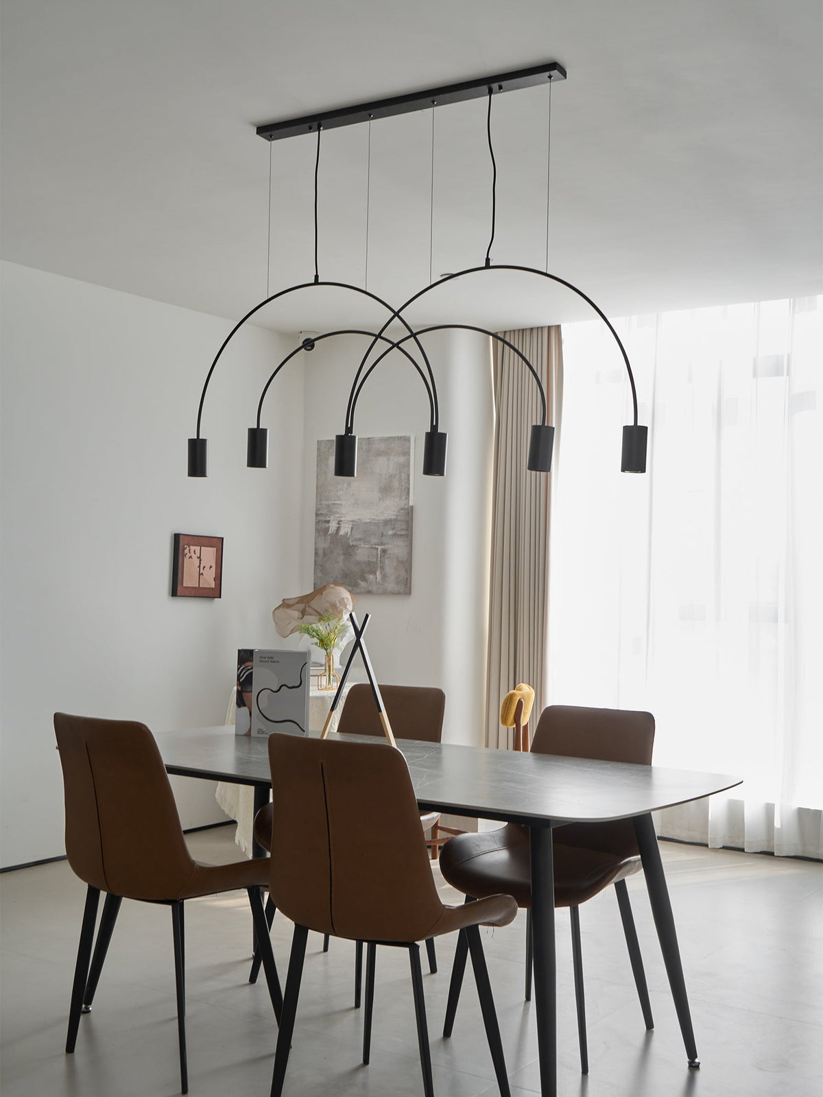 Tall view of a black Clérachic arch pendant above a round-cornered dark table, with brown chairs and sheer curtains beyond.