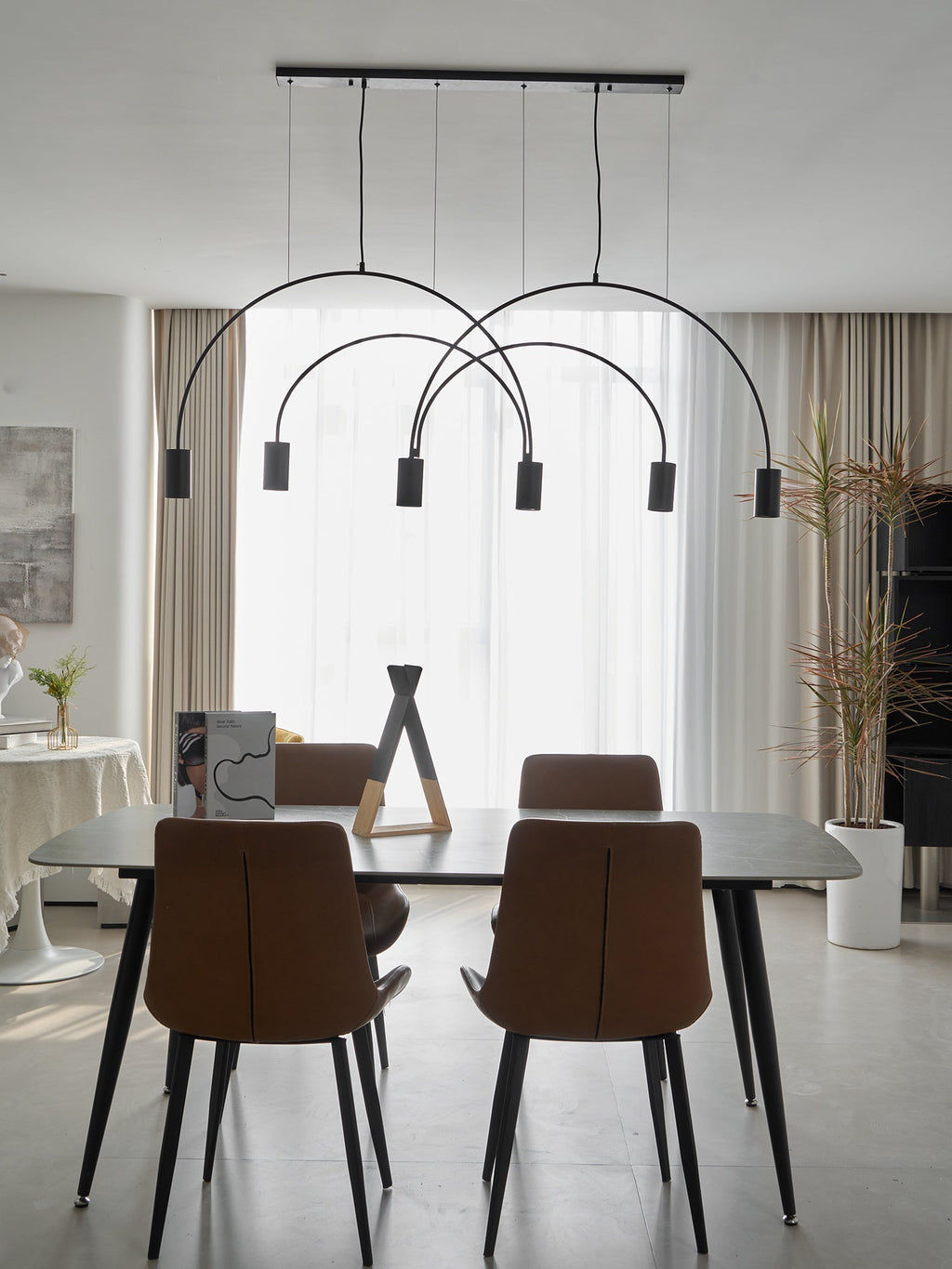 A black Clérachic arch pendant floats in front of linen curtains above a dining table, a tall dried-grass stem to the right.