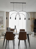 A black Clérachic arch pendant floats in front of linen curtains above a dining table, a tall dried-grass stem to the right.