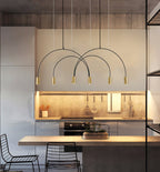 A brass-headed Clérachic arch pendant hangs in a grey kitchen with a lit worktop and two black wire chairs.