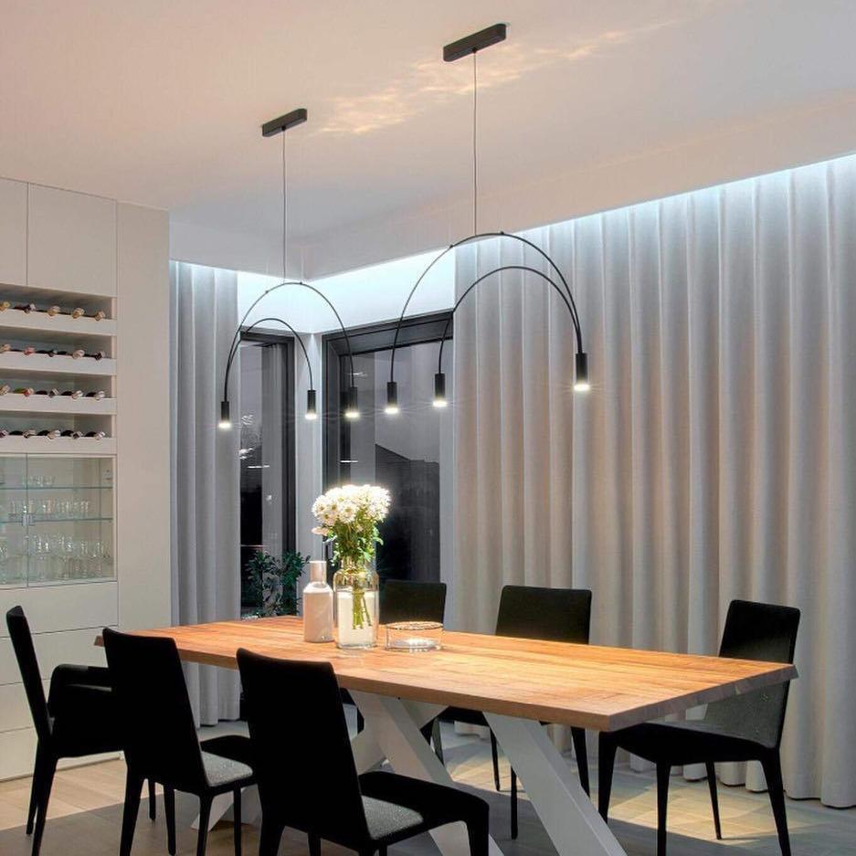 Two Clérachic arch pendants with slim black canopies hang over a wooden dining table with black chairs, in front of draped curtains.