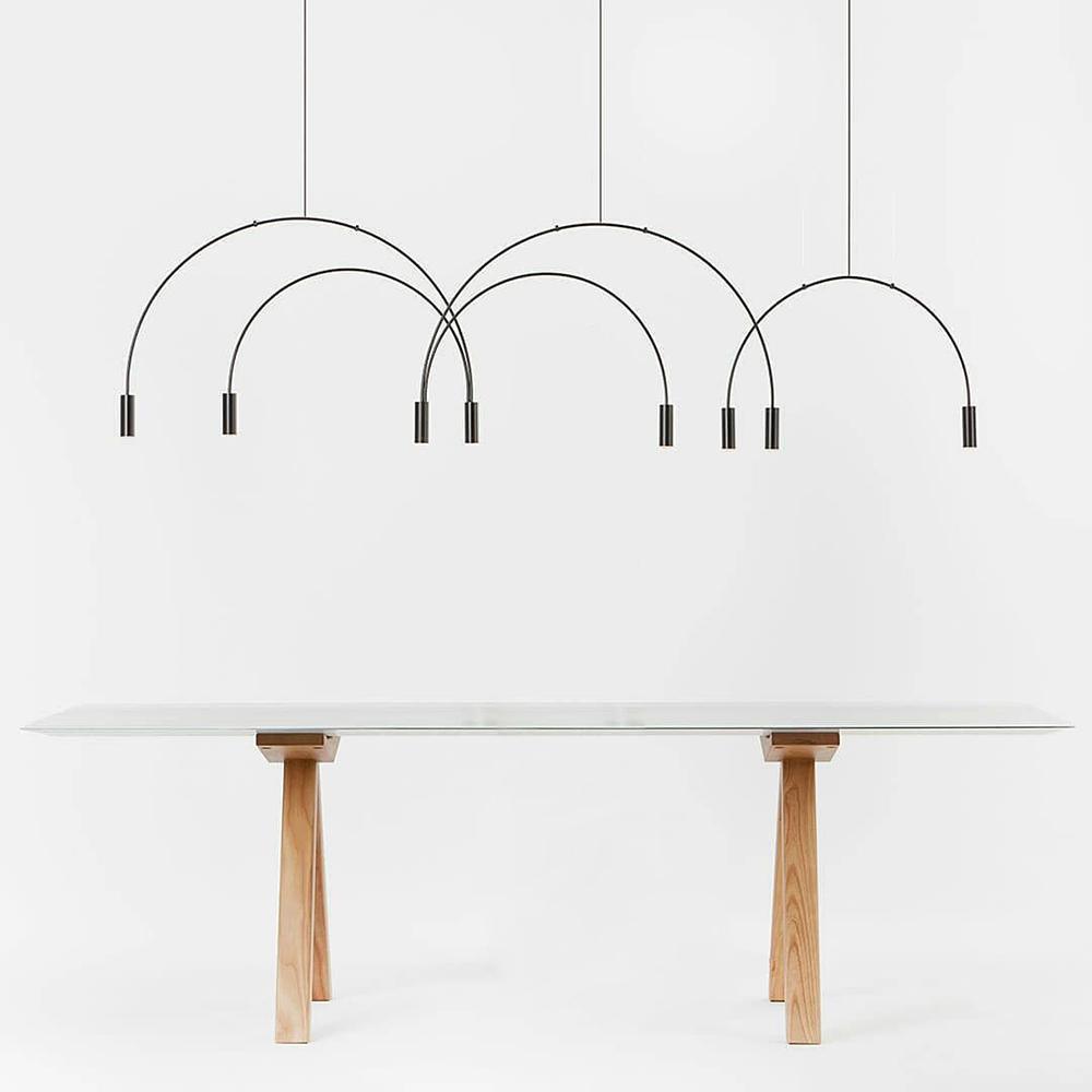 Eight slim black spot heads hang from this Clérachic arch pendant on a white backdrop above a table with timber trestle legs.