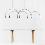 Eight slim black spot heads hang from this Clérachic arch pendant on a white backdrop above a table with timber trestle legs.