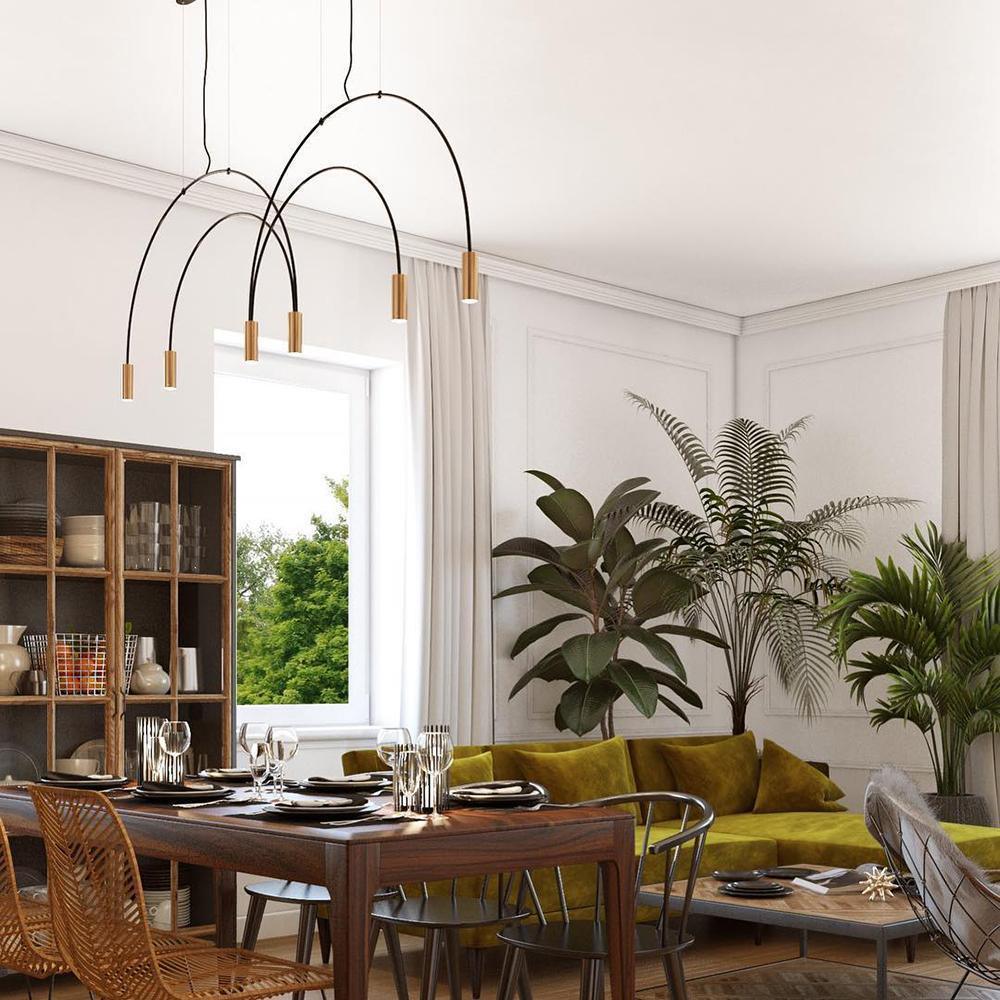 A Clérachic arch pendant with brass spot heads hangs in a dining room with an olive-green sofa, wicker chairs and palms.