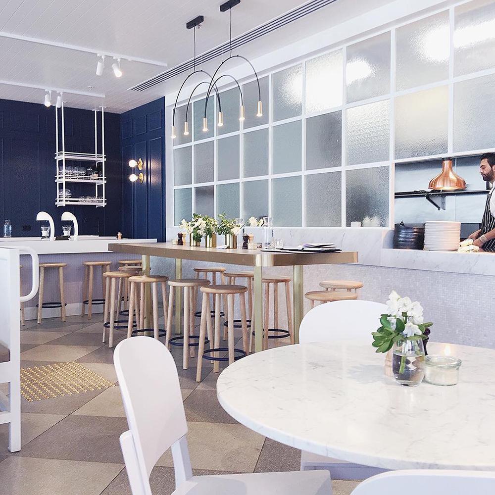 Two small Clérachic arch pendants hang over a long shared table in a bright cafe with white stools and navy panels.