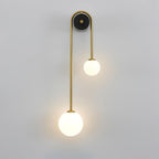 Clérachic arched brass wall sconce lit on a pale wall, the larger frosted globe hanging low on the long left leg.