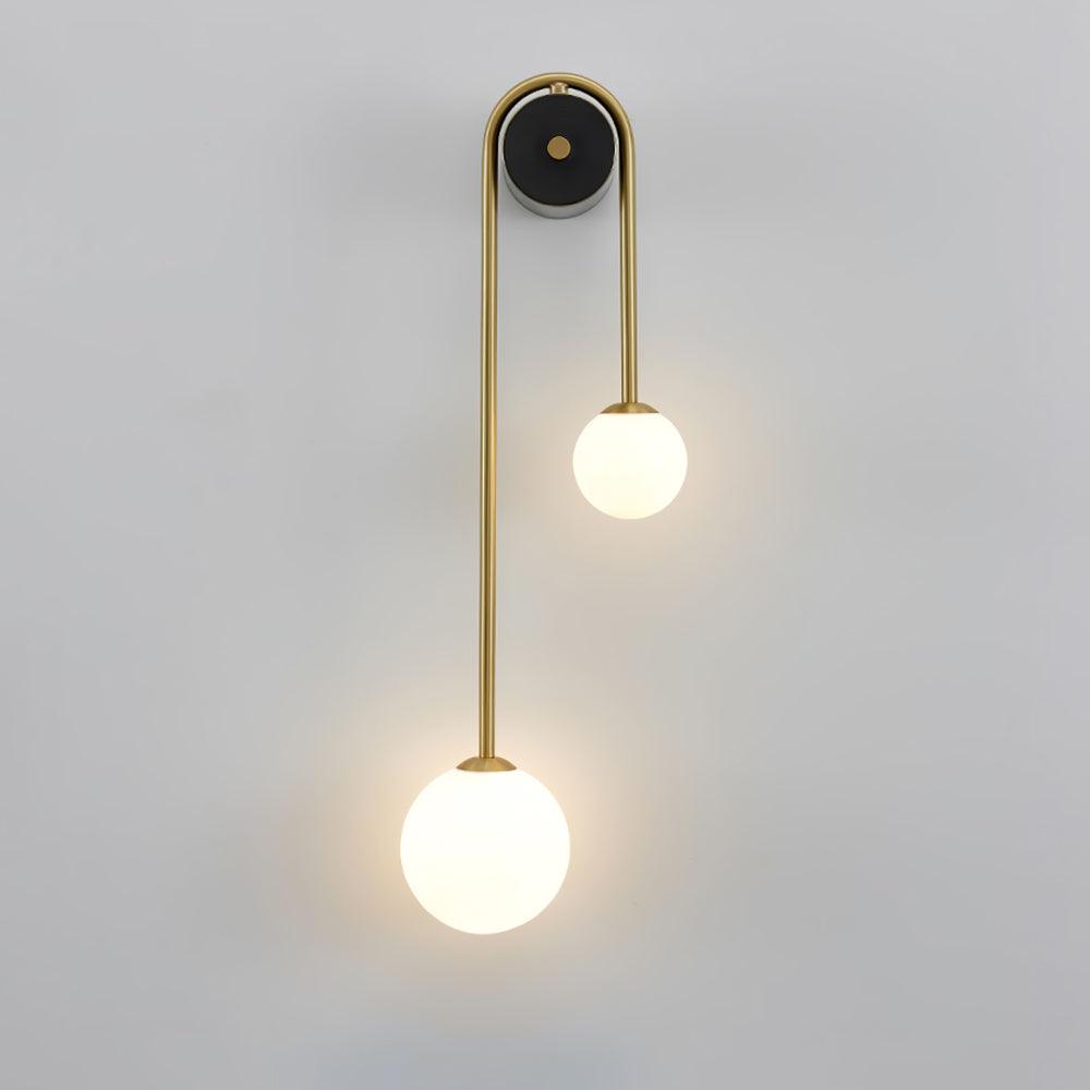 Clérachic arched brass wall sconce lit on a pale wall, the larger frosted globe hanging low on the long left leg.