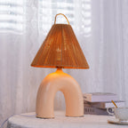 Close front view of an arched Skin ceramic Clérachic lamp glowing under its rope shade, on a draped table by sheer curtains.