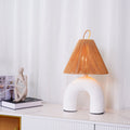 White arched ceramic Clérachic lamp with a rope shade on a white sideboard, beside a sculpted figure and a stack of books.