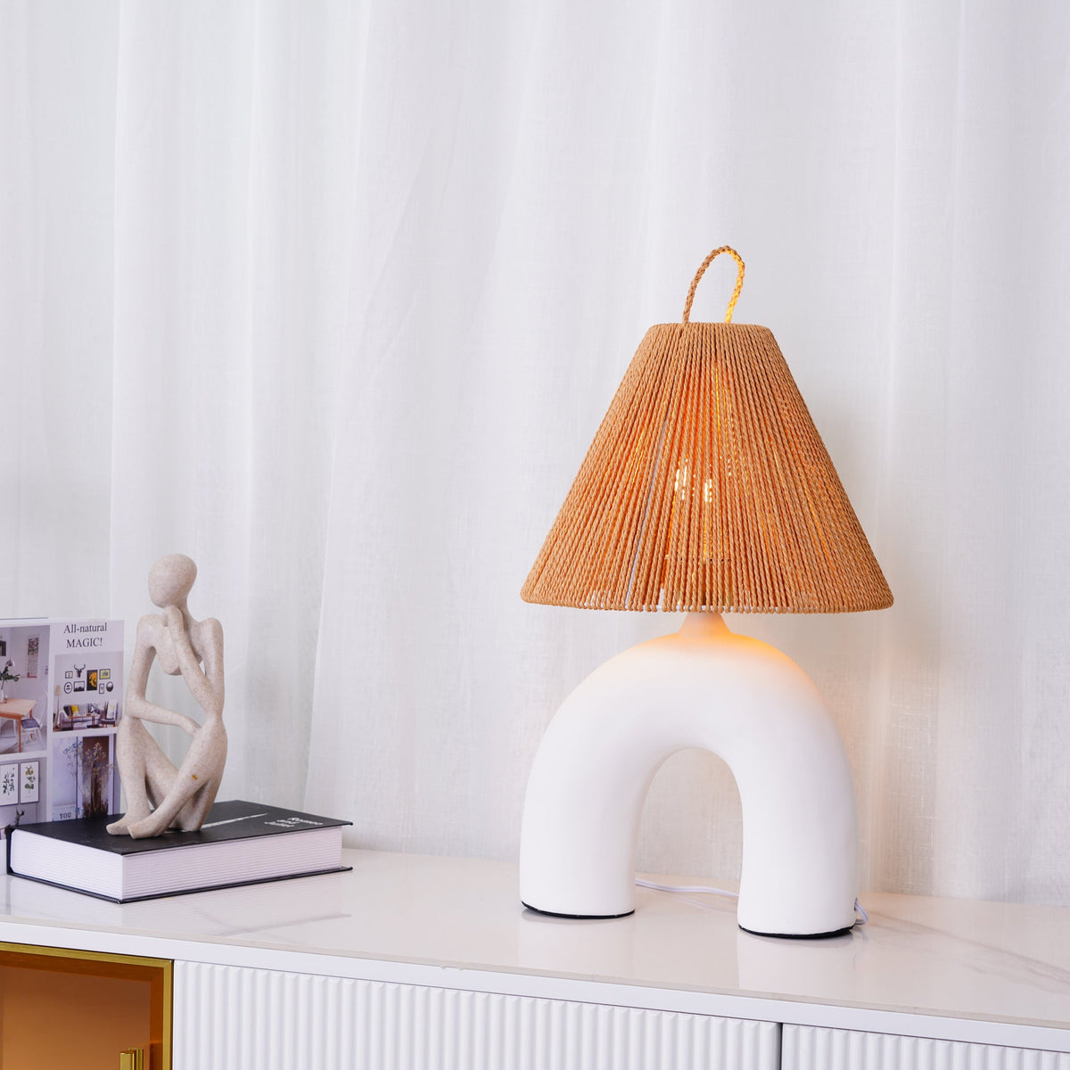 White arched ceramic Clérachic lamp with a rope shade on a white sideboard, beside a sculpted figure and a stack of books.