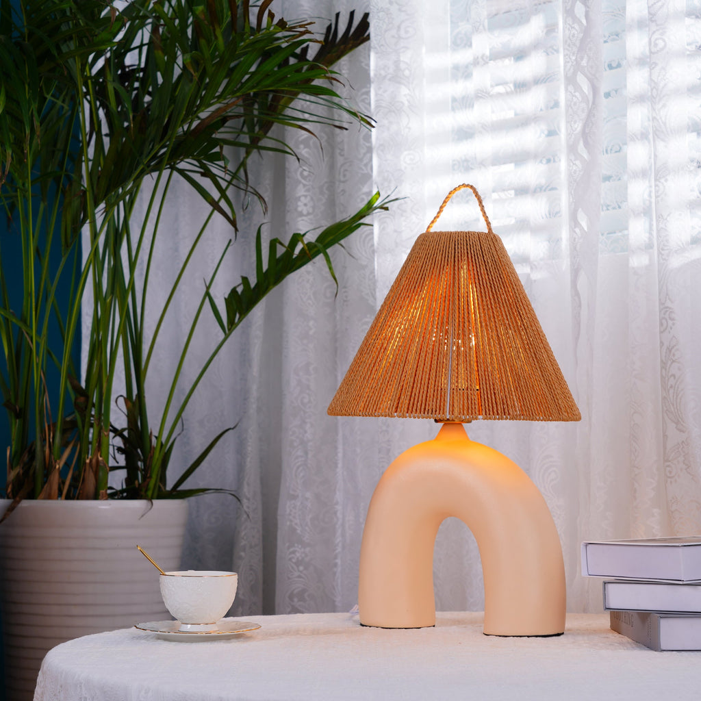 Lit Clérachic table lamp with a Skin arched base on a lace-covered table, with a tall palm, a teacup and books beside it.