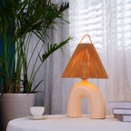 Lit Clérachic table lamp with a Skin arched base on a lace-covered table, with a tall palm, a teacup and books beside it.
