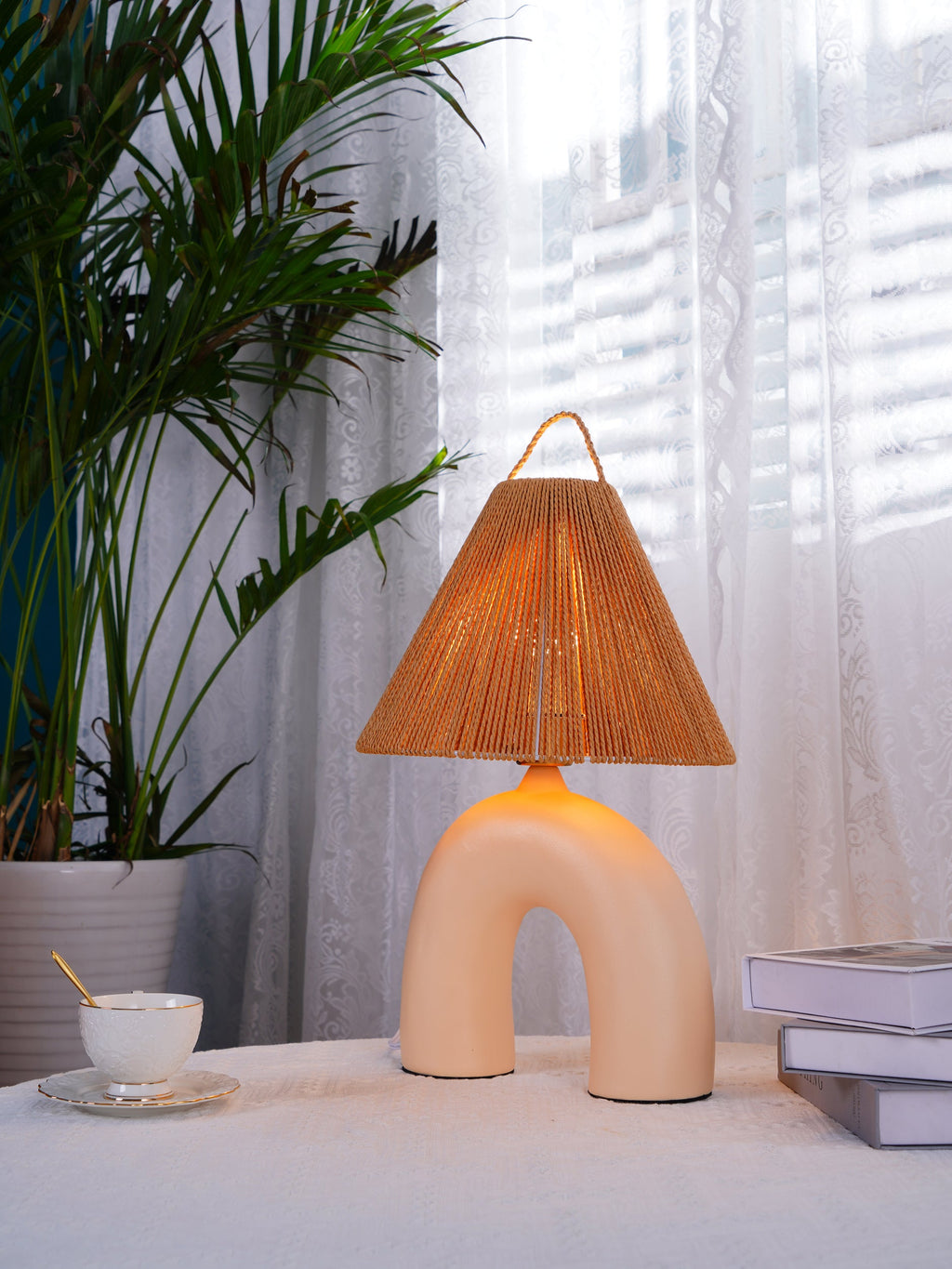 Narrower view of a Skin arched Clérachic lamp glowing on a round table, a leafy palm and sheer curtains behind it.
