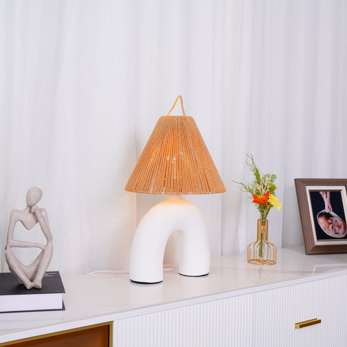 White arched Clérachic table lamp lit on a marble-topped sideboard, with a figurine, a small flower vase and a framed photo.