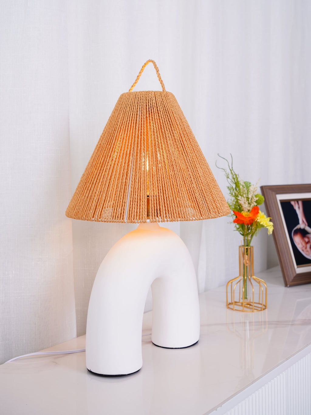 Close view of a white arched ceramic Clérachic lamp under a woven rope shade, with a bud vase and framed photo beside it.