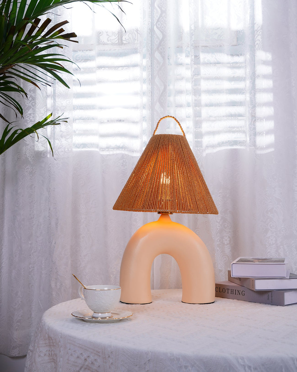 Skin arched Clérachic table lamp lit on a lace table cloth, with a teacup and saucer at its foot and palm leaves behind.
