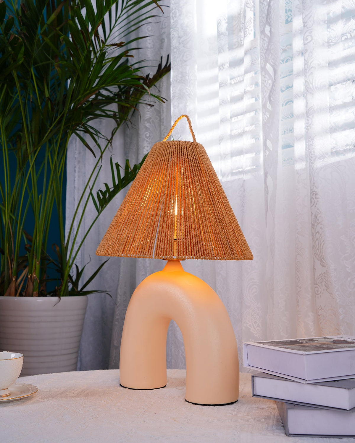 Side view of this Clérachic lamp with a Skin arched base and glowing rope shade, beside a stack of books and a palm.