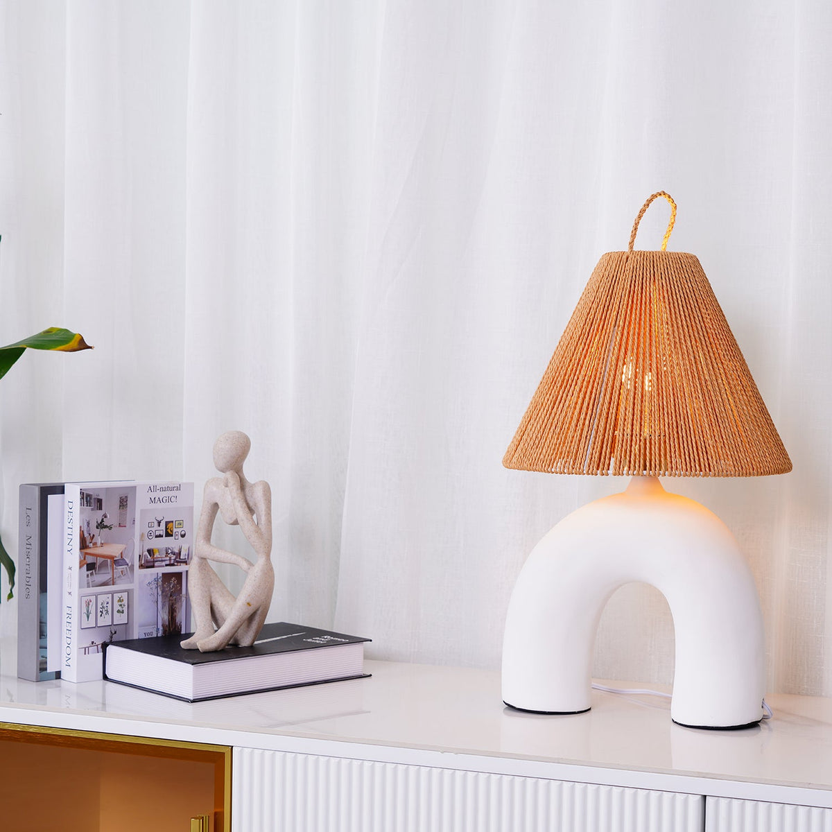 Wide view of a white arched Clérachic lamp on a white sideboard with gold handles, with a figurine and stacked books.